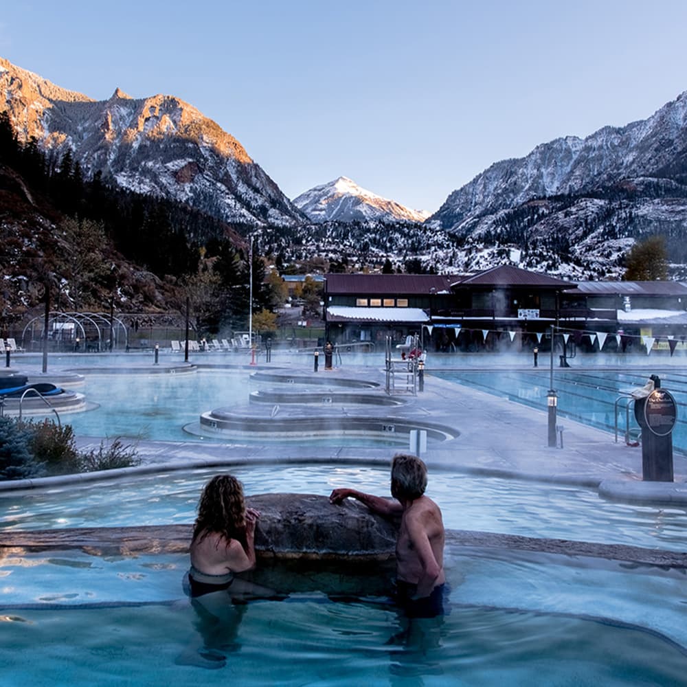 Two people relax in a steaming hot spring pool with snowy mountains in the background.