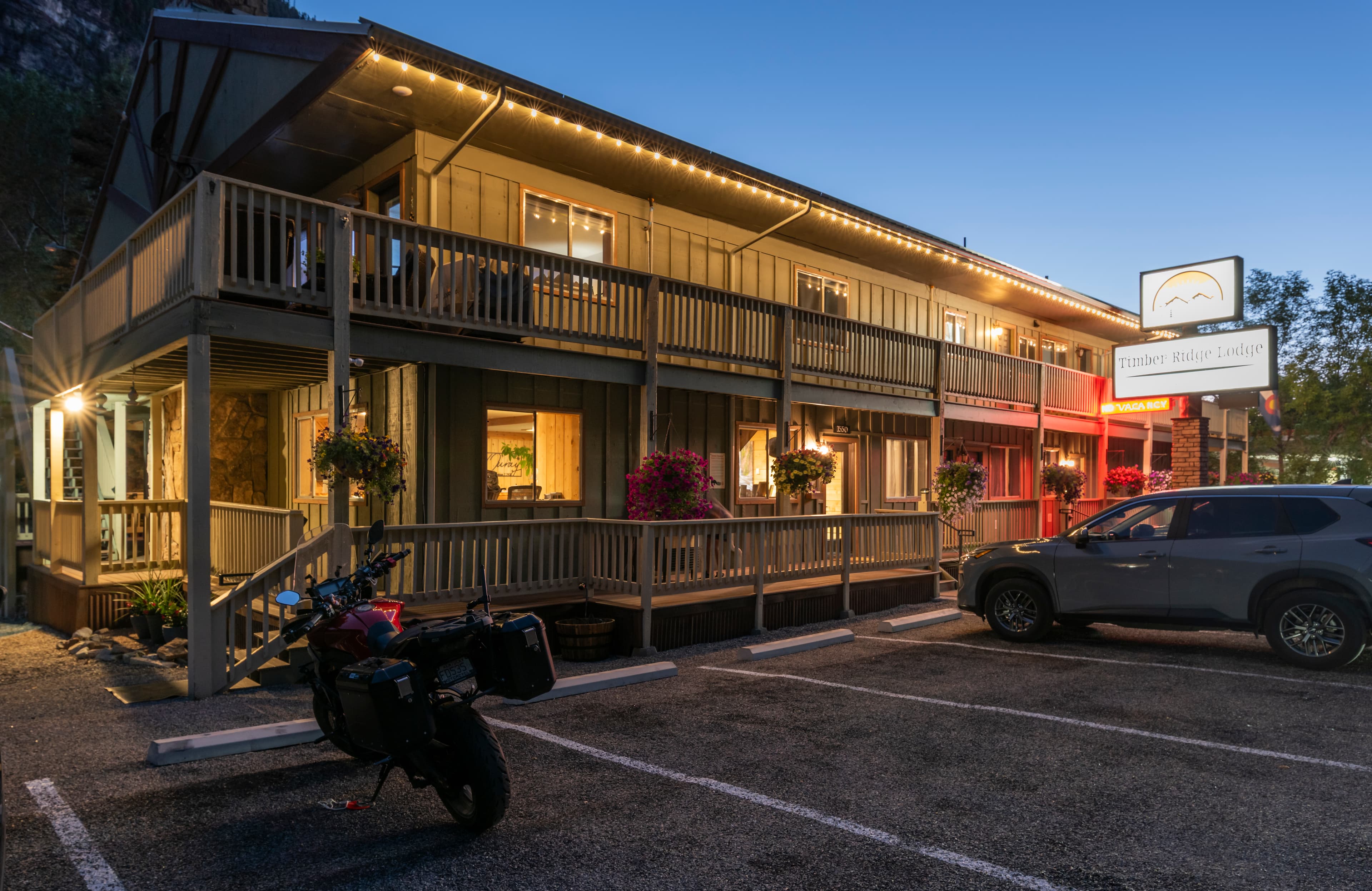 A wooden motel building illuminated at dusk, with a sign and parked vehicles nearby.