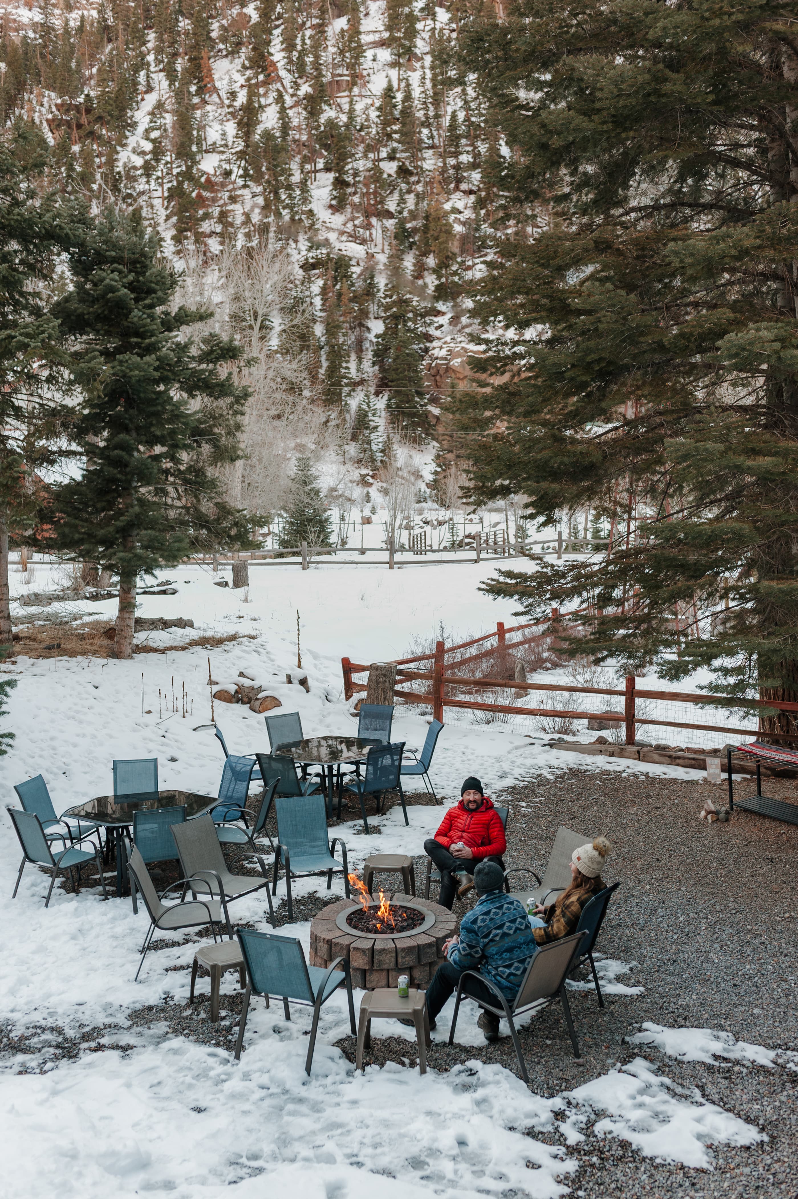 Three people sit around a fire pit in a snowy landscape with scattered chairs and trees.