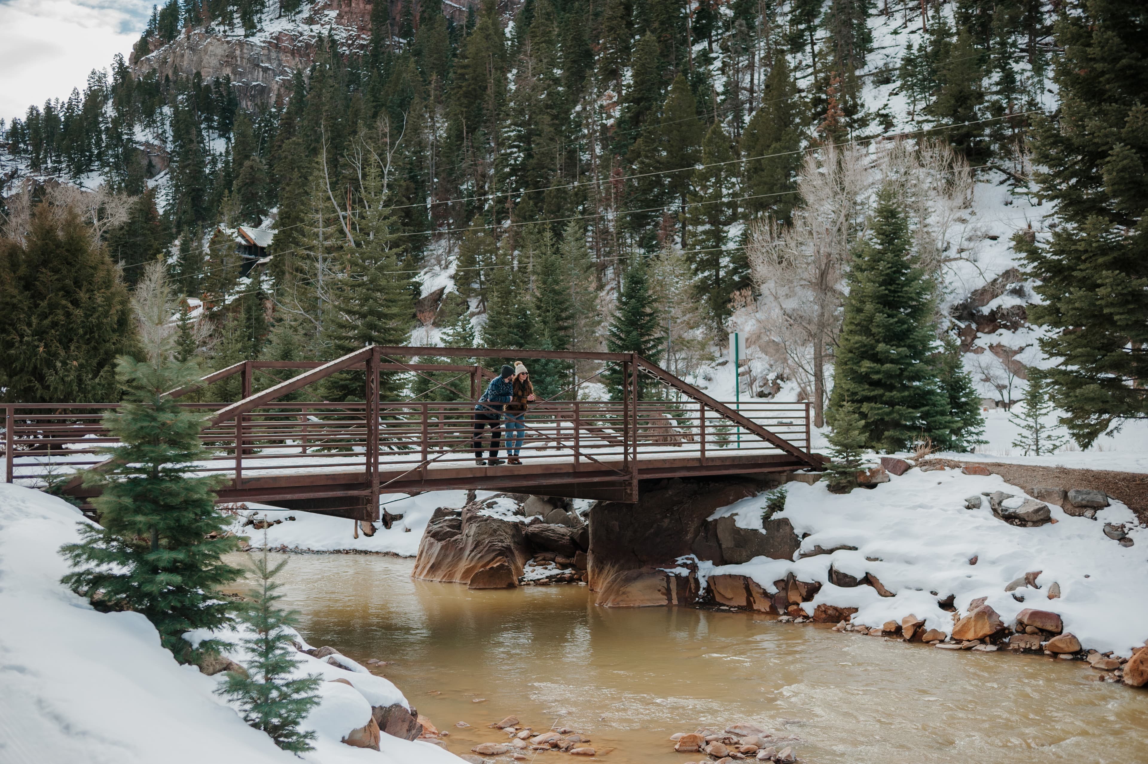 A couple embraces on a wooden bridge over a snow-covered stream surrounded by evergreen trees.