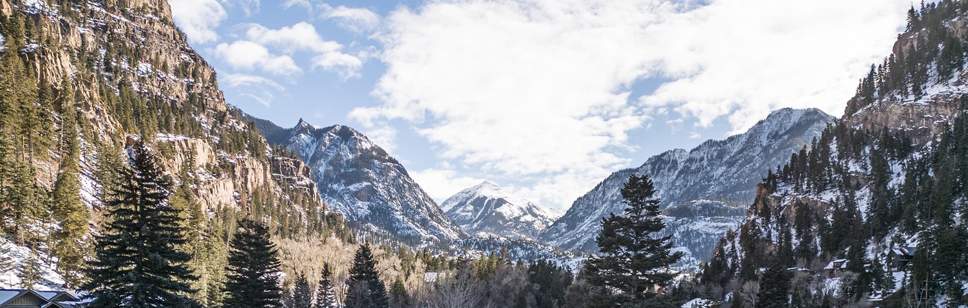 A scenic view of snow-capped mountains surrounded by trees under a partly cloudy sky.