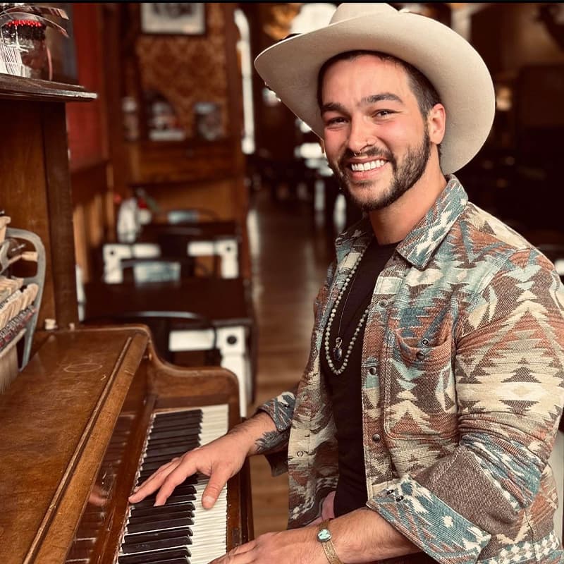A smiling man in a cowboy hat plays a piano in a cozy, rustic setting.