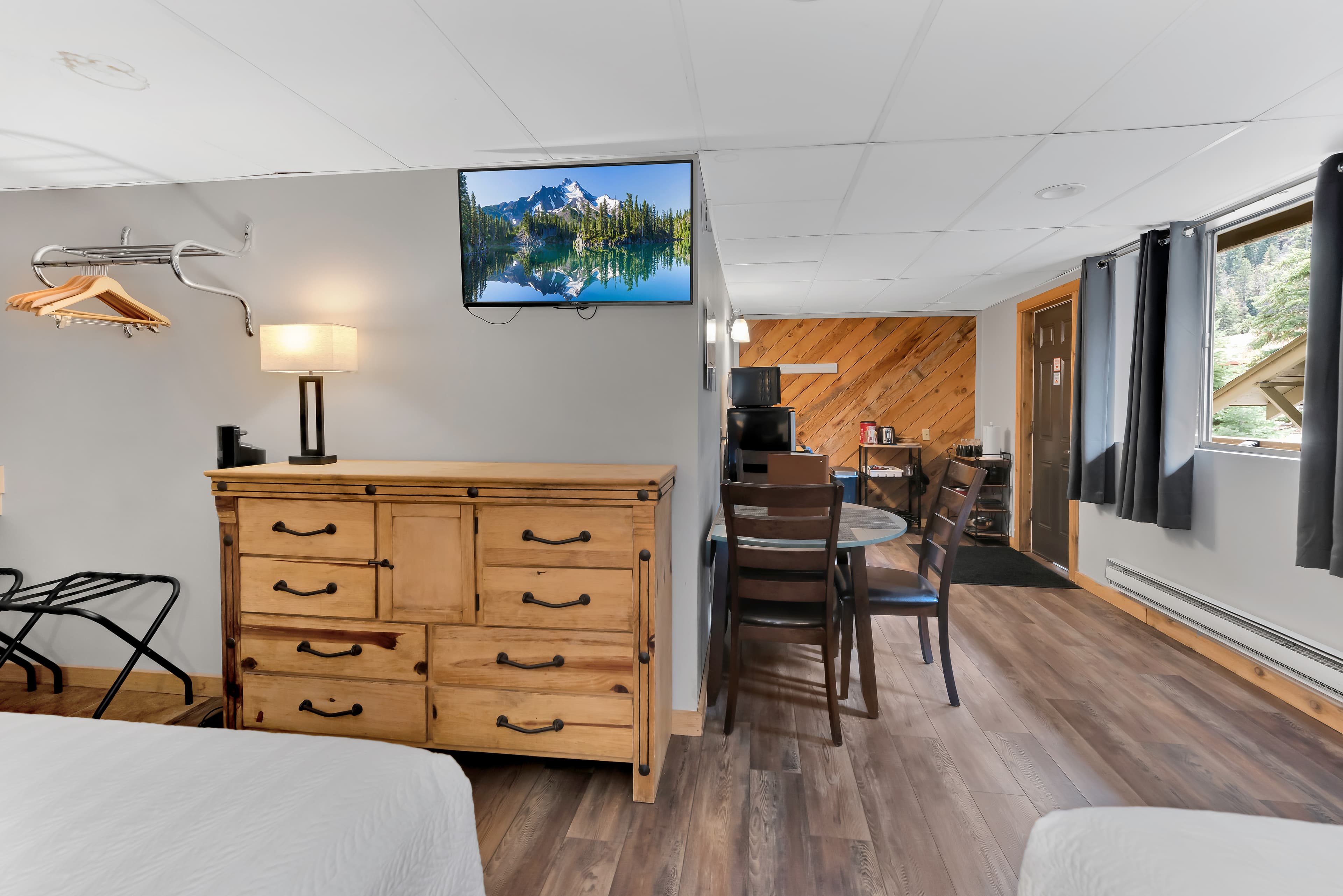 A view from the sleeping area looking toward the kitchenette. In the foreground is a large light-wood dresser with a flat-screen TV mounted above it. Beyond the dresser, there is a circular dining table for two and a glimpse of the kitchen area with a wood-paneled accent wall.