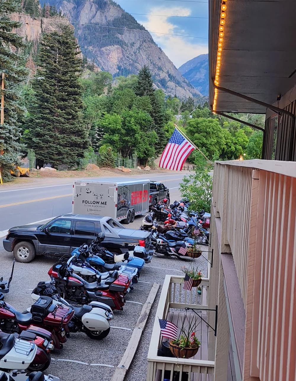 A view from a balcony showing parked motorcycles, a pickup truck, and a large American flag amidst mountainous surroundings.