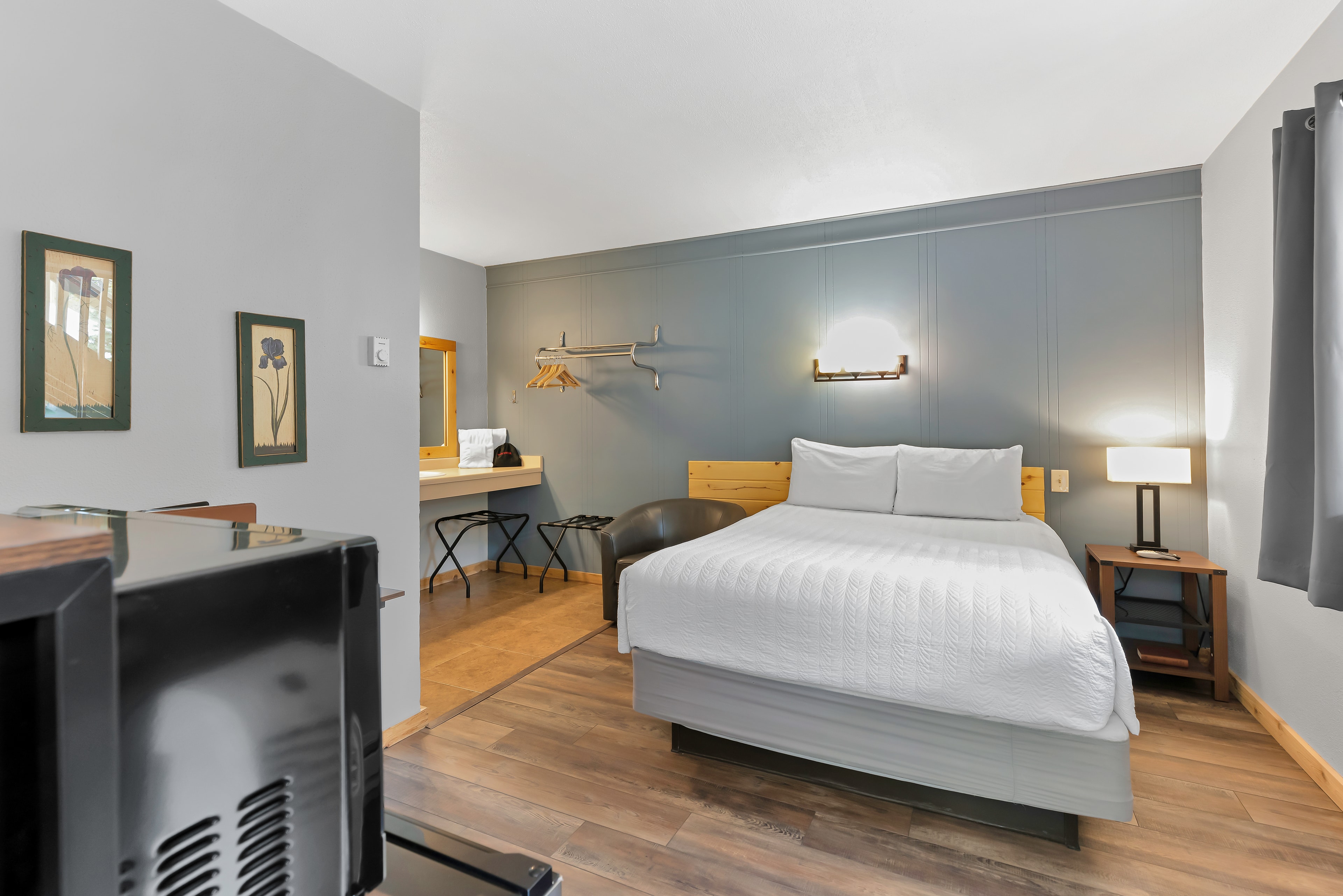 A welcoming queen guest room at Timber Ridge Lodge featuring a plush queen bed with a wood-slat headboard. The room showcases a sage green accent wall, wood-style flooring, and a convenient vanity area in the background, offering a cozy retreat for Ouray visitors.
