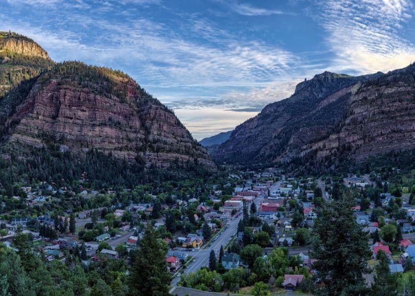 A panoramic view of a valley surrounded by mountains, featuring a small town below and a blue sky with wispy clouds.
