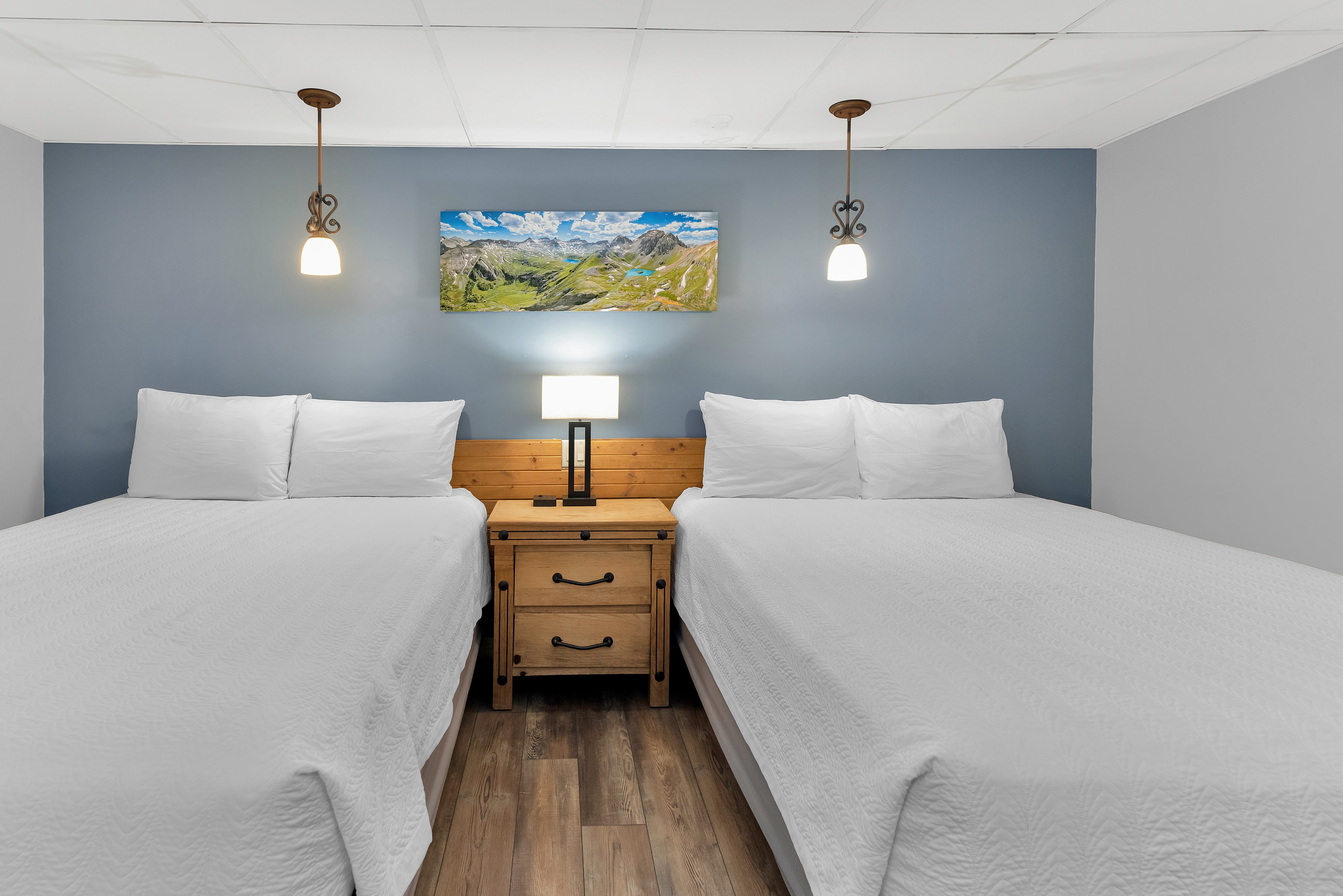 A symmetrical view of a guest room at Timber Ridge Lodge featuring two queen beds with crisp white linens. A rustic wooden nightstand sits between the beds with a modern lamp, set against a blue accent wall decorated with a panoramic mountain landscape photo.