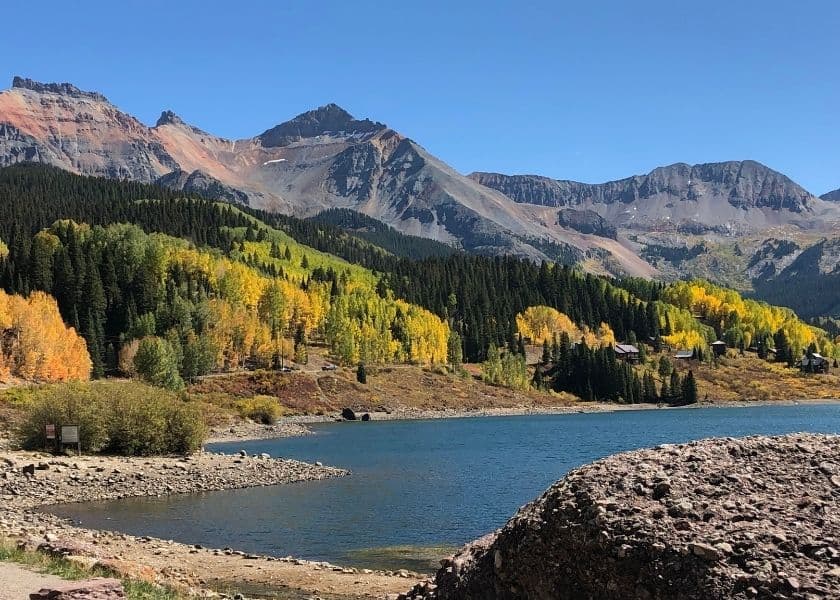 Trout Lake with blue water, rocky shoreline, golden fall trees, evergreen forest, and rugged mountain peaks.